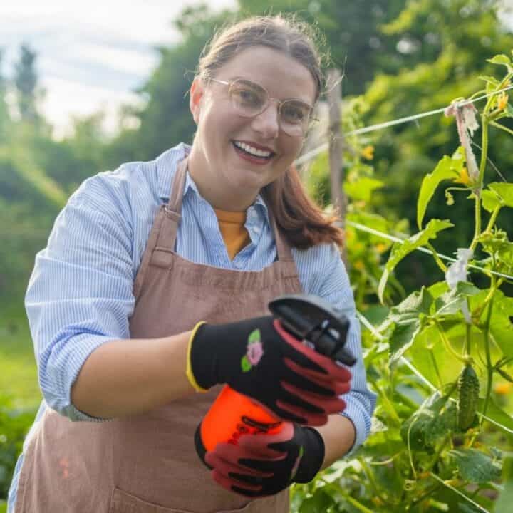A woman in an apron and gloves smiles while holding a spray bottle and tending to plants in a garden.