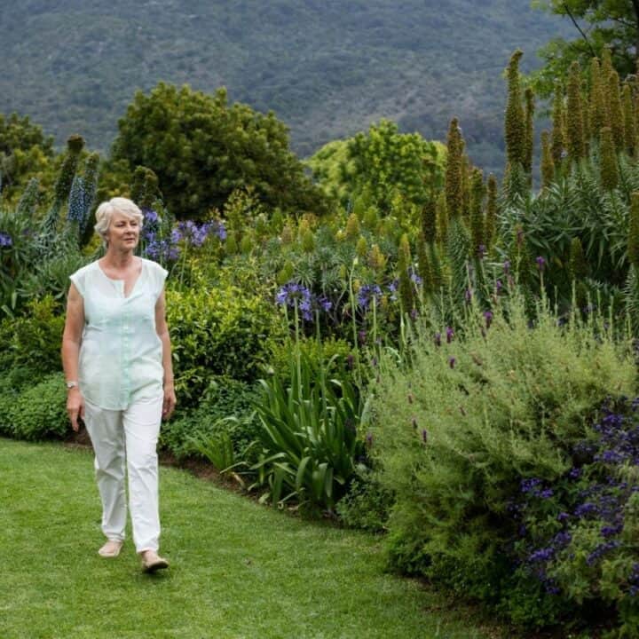 An older woman in light clothing walks along a grassy path surrounded by lush green plants and flowers in a garden.