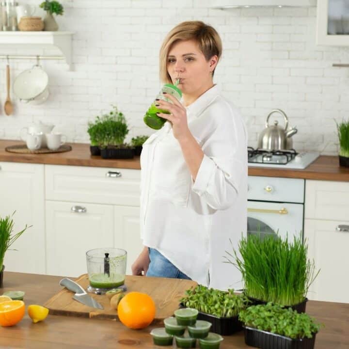 A woman stands in a kitchen drinking a green smoothie, surrounded by fresh greens, citrus fruits, and a blender on the counter.
