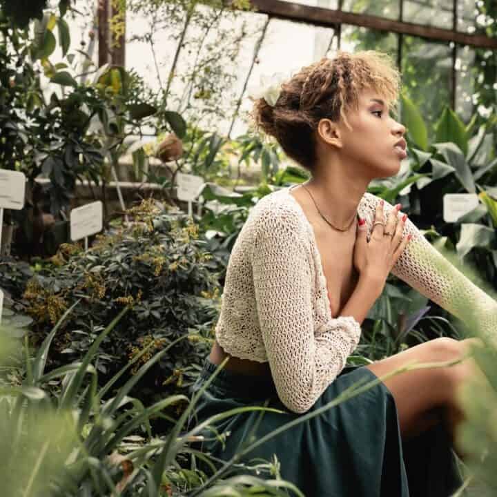 A woman sits on a bench in a greenhouse surrounded by green plants, wearing a cream knit top and dark skirt, looking to the side with one hand on her chest.