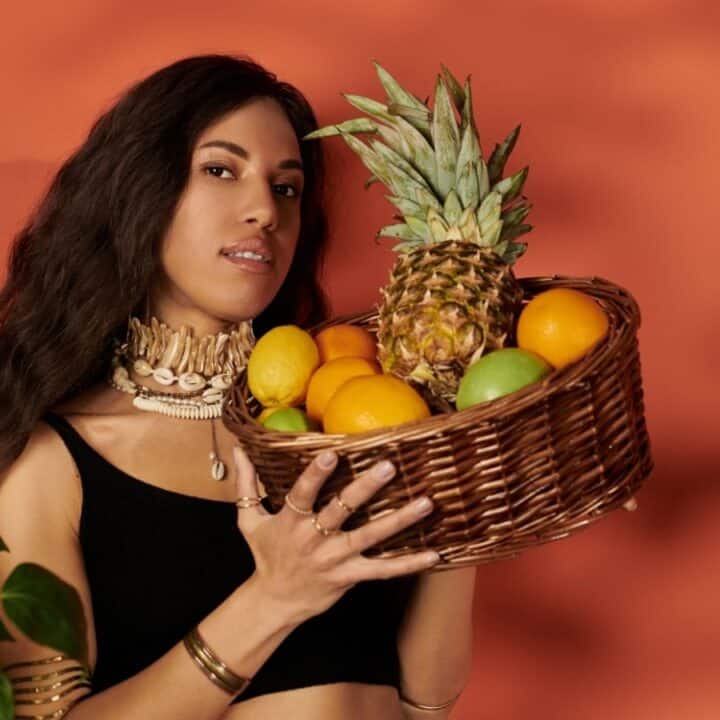 A woman wearing a black top and shell necklace holds a wicker basket filled with a pineapple, oranges, and other fruits against an orange background.