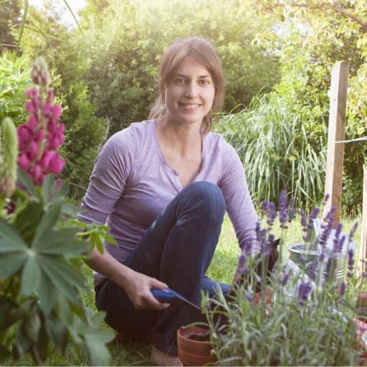A woman kneels in a garden surrounded by green plants and flowers, holding gardening shears and smiling at the camera.