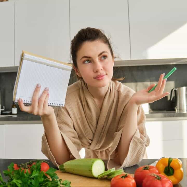 A woman in a kitchen holds a notepad and pen, surrounded by vegetables and cooking ingredients on the counter.