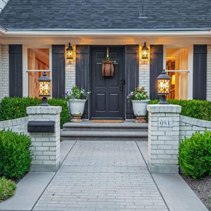 Front entryway of a brick house with black door, potted plants, lantern-style lights, trimmed hedges, and a "One" address sign on the right brick pillar.