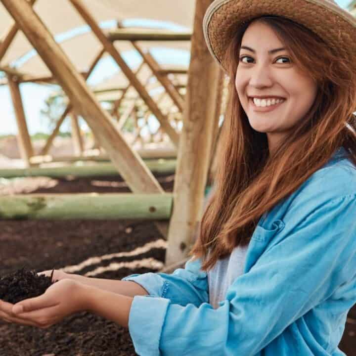 A woman in a straw hat and blue shirt holds soil in her hands and smiles in a covered outdoor structure.