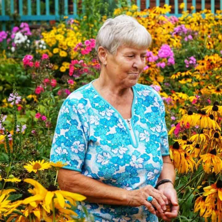 An elderly woman with short gray hair stands in a garden surrounded by blooming yellow and pink flowers, wearing a blue floral-patterned shirt.