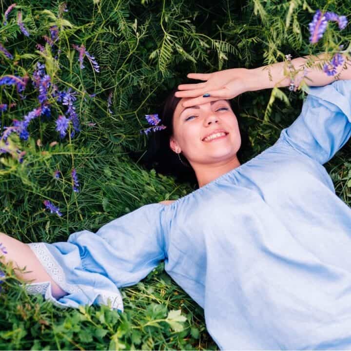 A woman in a light blue dress lies on green grass surrounded by purple wildflowers, smiling and shielding her eyes from the sun with one hand.