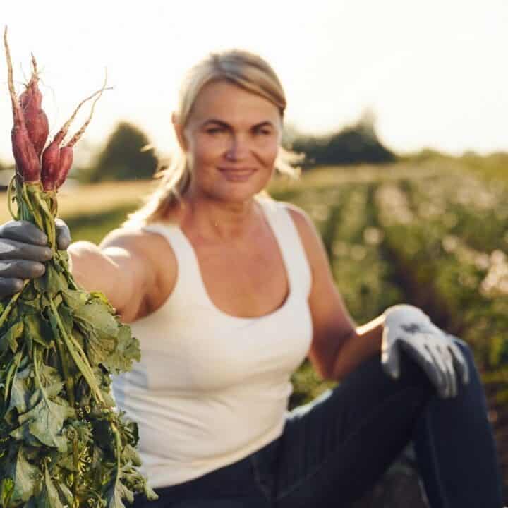 A woman in a white tank top and gloves holds up freshly harvested radishes while kneeling in a vegetable field.