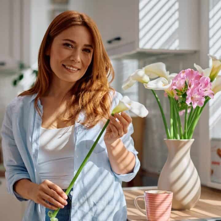 A woman stands in a sunlit kitchen holding a white flower, with a vase of flowers and a striped mug on the counter beside her.