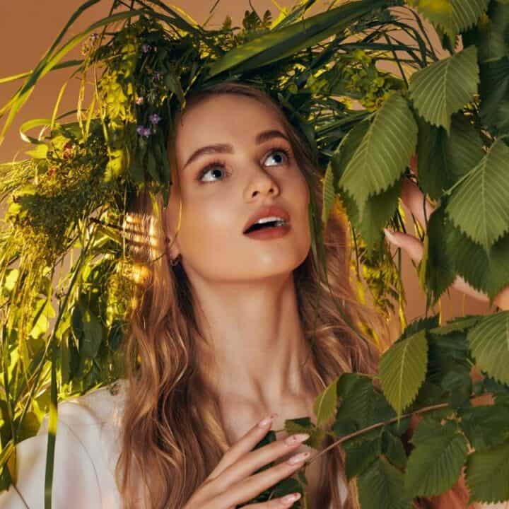 A woman with long, wavy hair wears a headdress made of green plants and leaves, surrounded by foliage, looking upward with a neutral expression.
