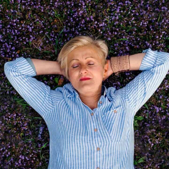 A person with short blonde hair lies on their back in a patch of purple flowers, eyes closed and arms behind their head, wearing a blue and white striped shirt.