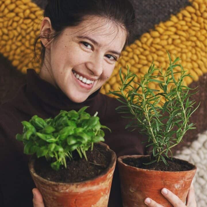 A person smiles while holding two potted plants, one basil and one rosemary, standing in front of a textured, patterned background.