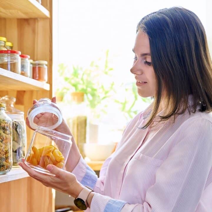 A woman in a light pink shirt takes dried fruit from a glass jar in a pantry with shelves stocked with jars of food.