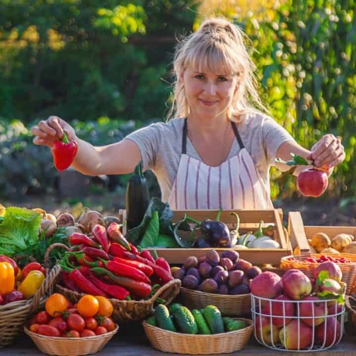 A woman stands behind a table covered with various fresh fruits and vegetables in an outdoor garden setting, holding a red bell pepper and an apple.
