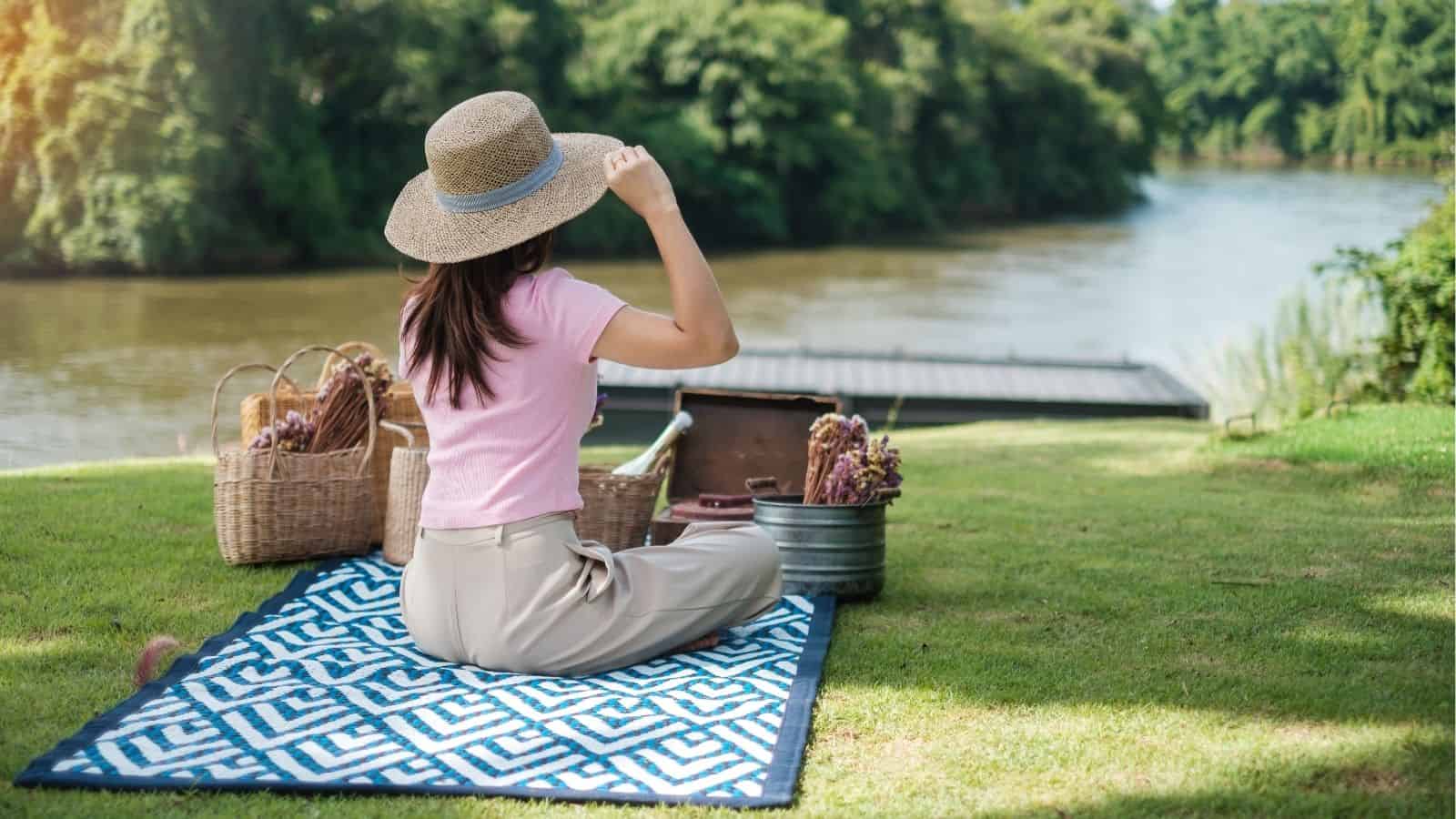 A person wearing a hat and pink shirt sits on a patterned mat by a river, with picnic baskets and food beside them on a grassy area.