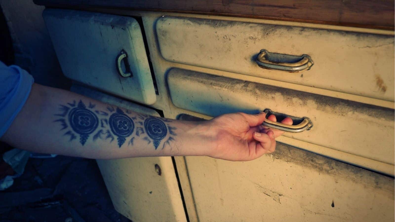 A person with a sun and circle tattoo on their forearm is opening a stained, old metal drawer with a brass handle.