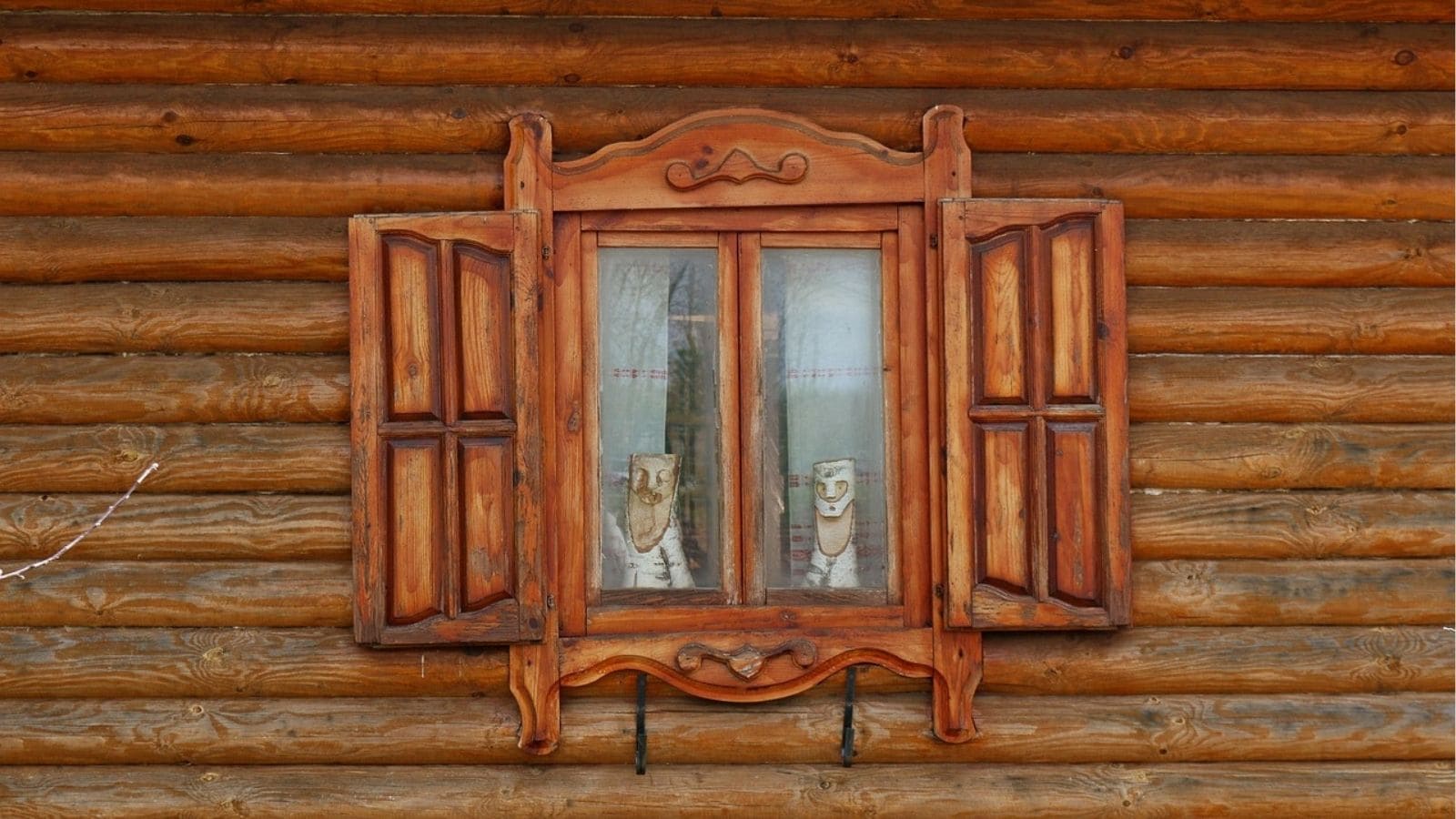 Wooden-framed window with open shutters set in a log cabin wall; lace curtains and two decorative figures are visible inside.