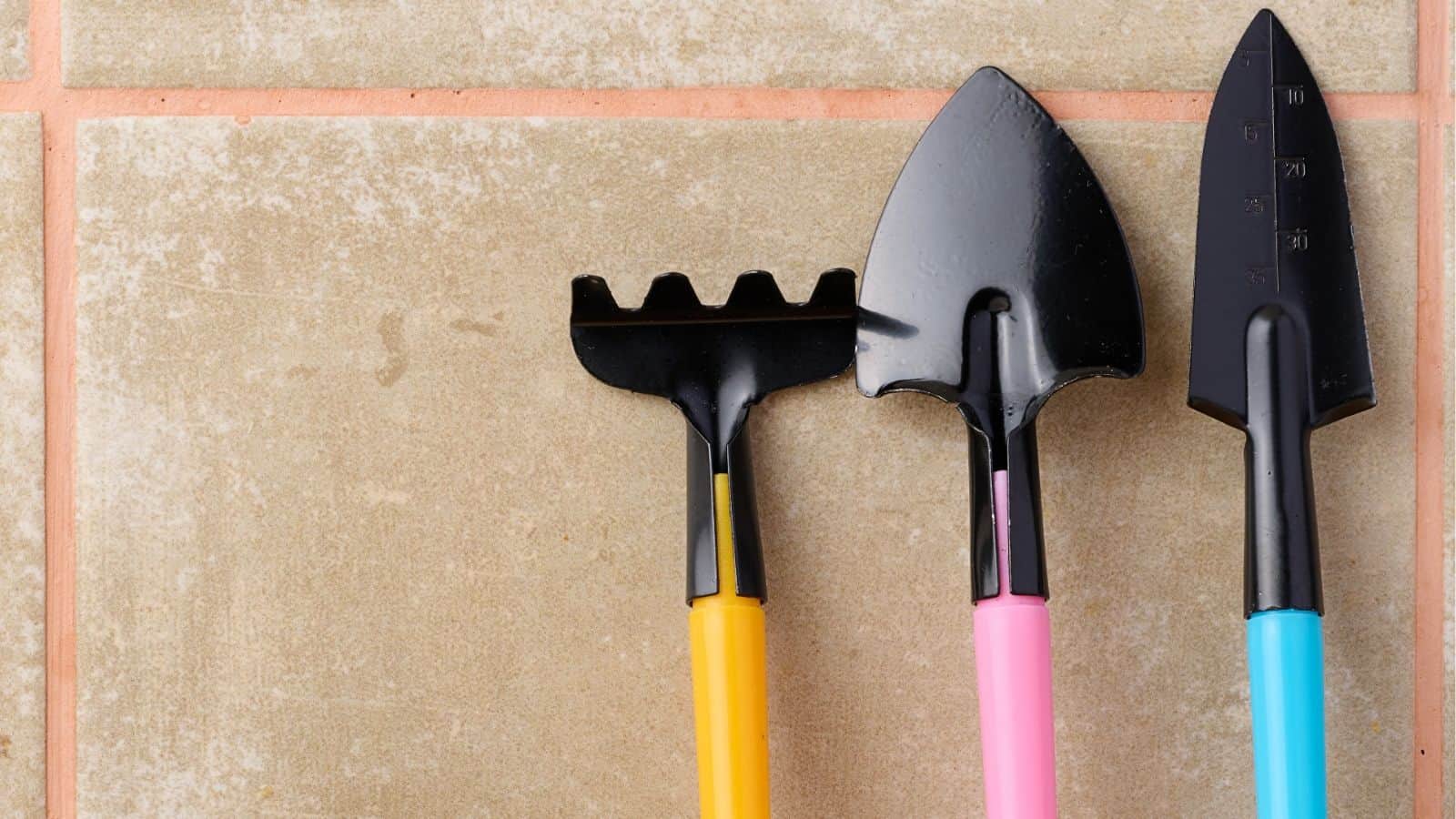 Three small gardening tools with colored handles (yellow, pink, blue) are arranged on a tiled surface. The tools include a rake, a spade, and a pointed trowel.