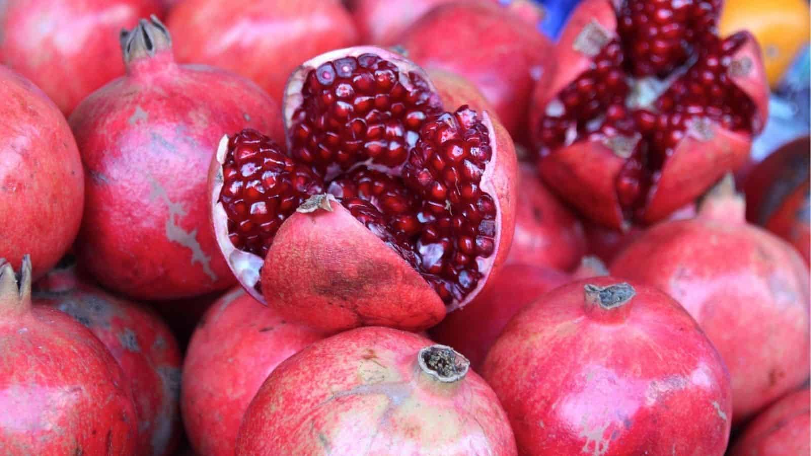 A pile of whole pomegranates with two split open, revealing clusters of red seeds inside.