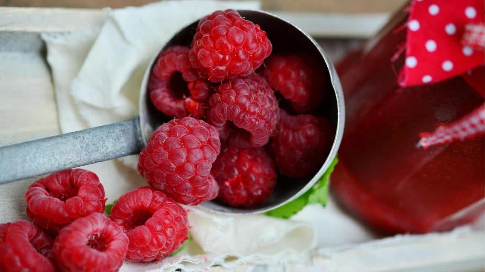 A metal measuring cup filled with fresh raspberries sits on a white cloth, with more raspberries nearby and a jar of raspberry jam to the side.