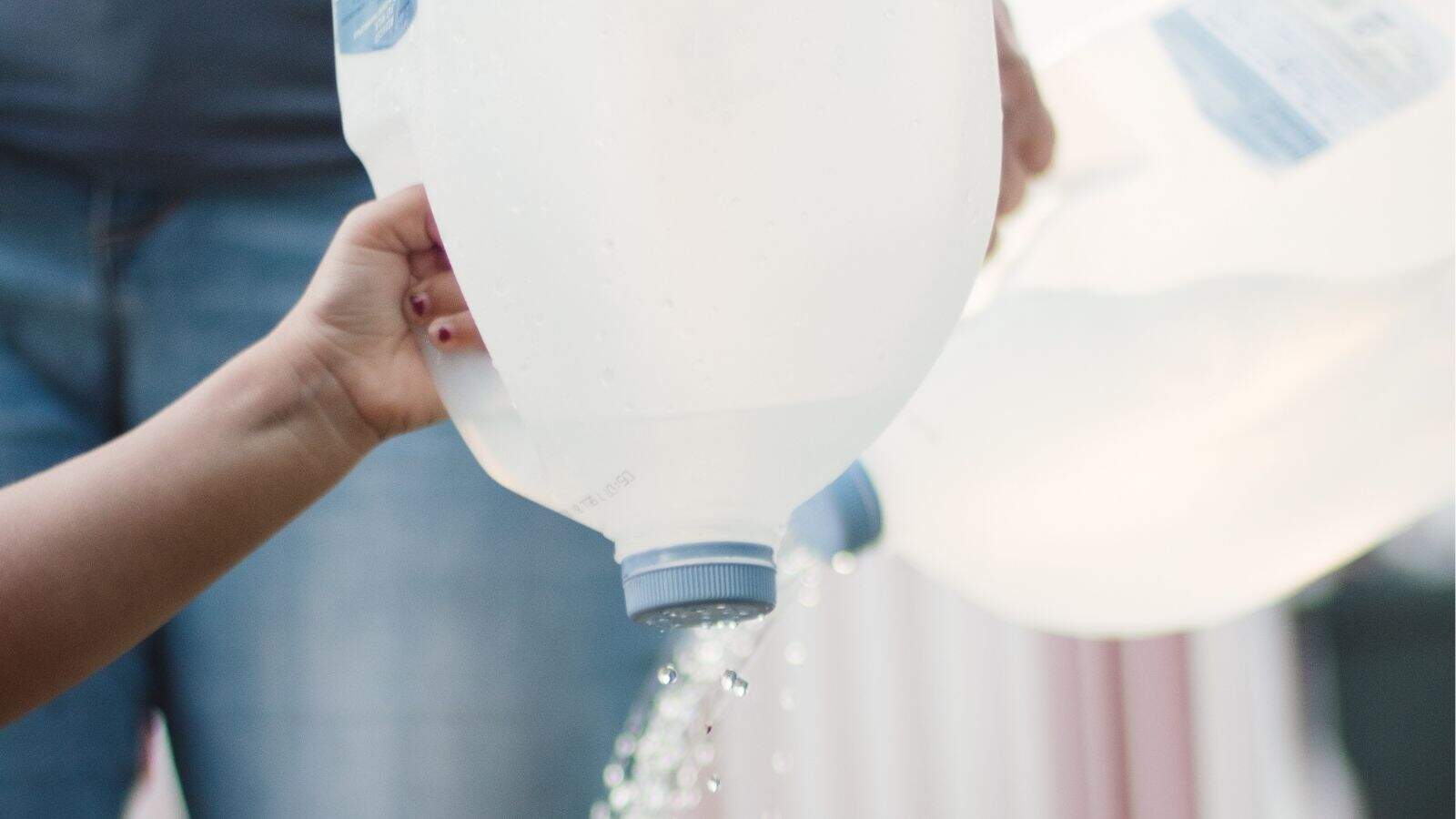 A hand holds an upturned plastic water jug with water pouring out, while another person in jeans holds a similar jug in the background.