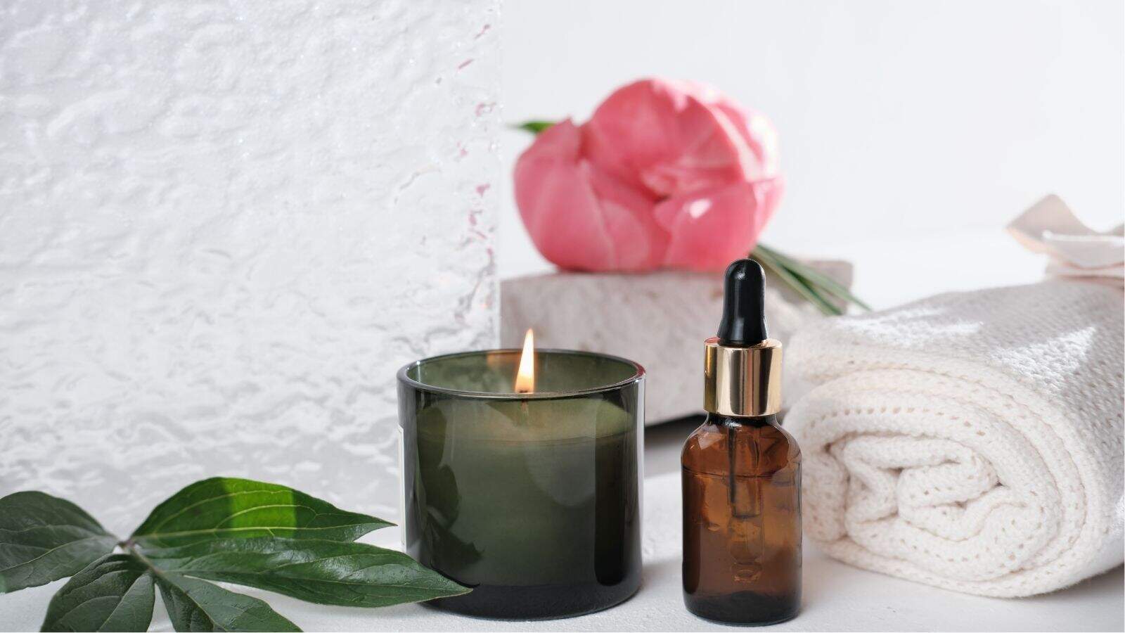 A lit green candle, a brown dropper bottle, a folded white towel, a pink flower, and green leaves arranged on a white surface.