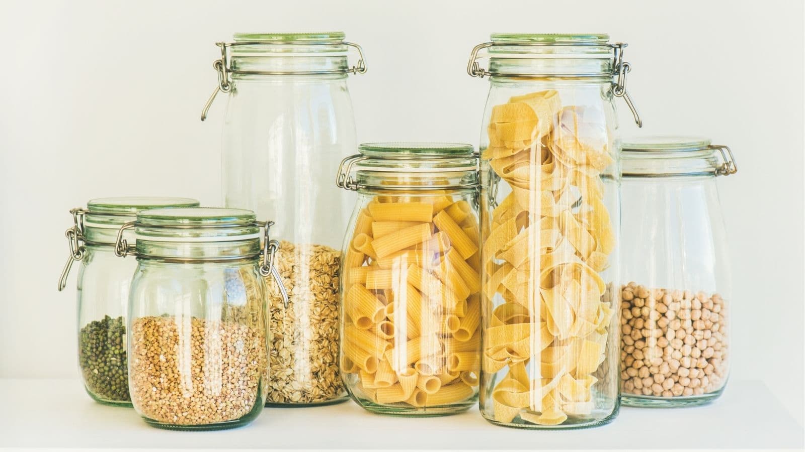 Six glass jars filled with various dry pantry staples, including green lentils, oats, rigatoni pasta, shell pasta, and chickpeas, arranged on a white surface against a light background.