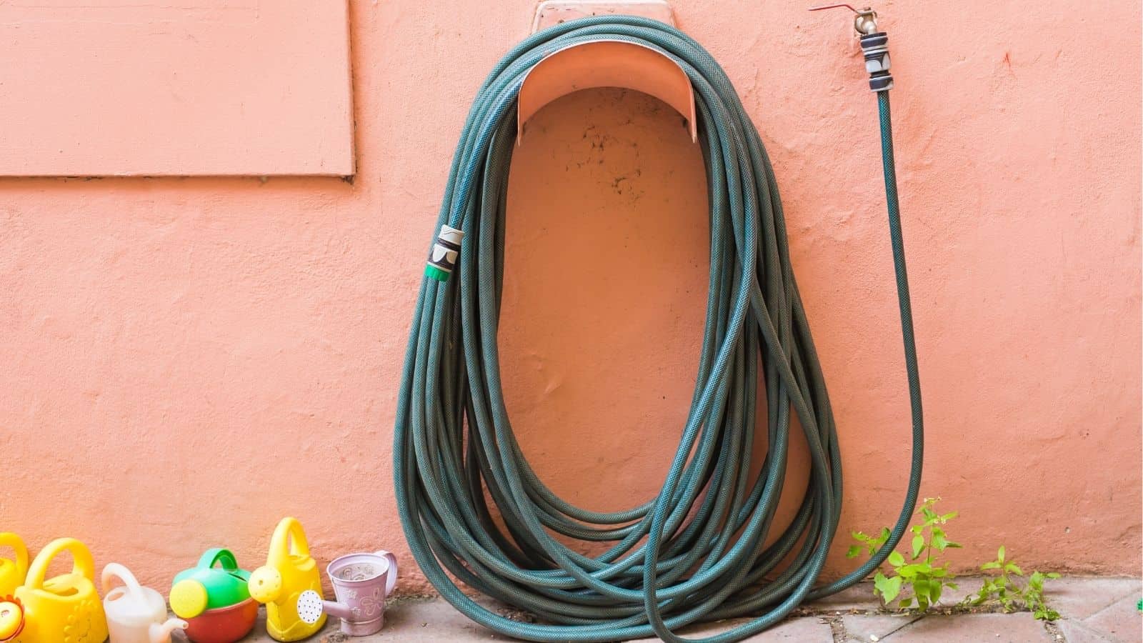 A coiled garden hose is mounted on a peach-colored wall with several small watering cans and a potted plant on the ground nearby.