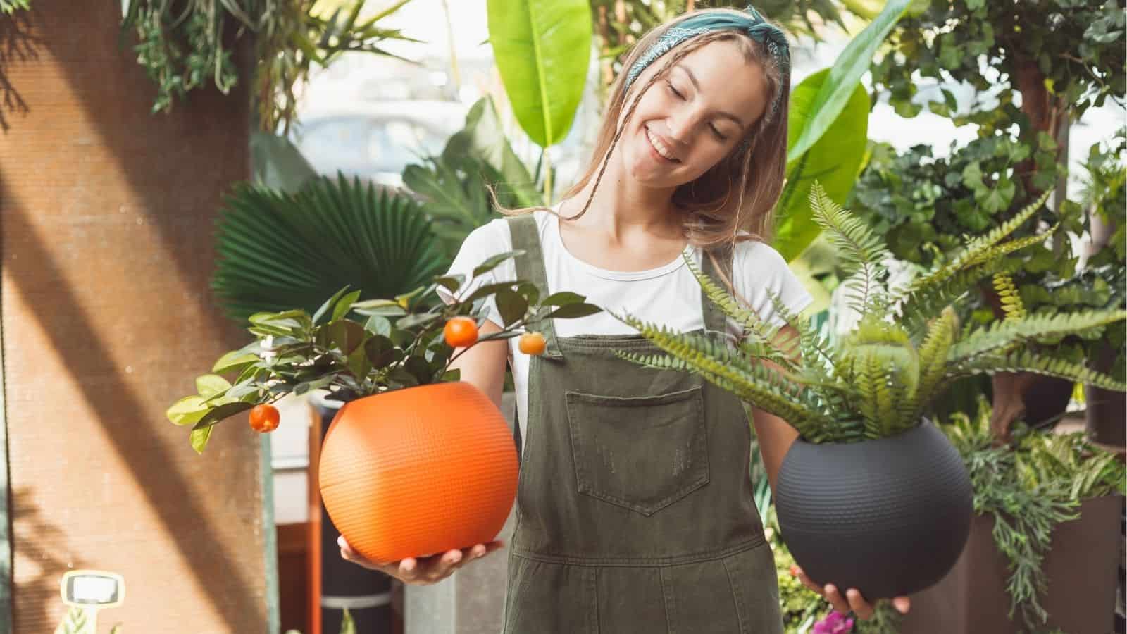 A woman in green overalls and a white shirt is holding two potted plants, one in an orange pot and the other in a black pot, surrounded by various greenery.