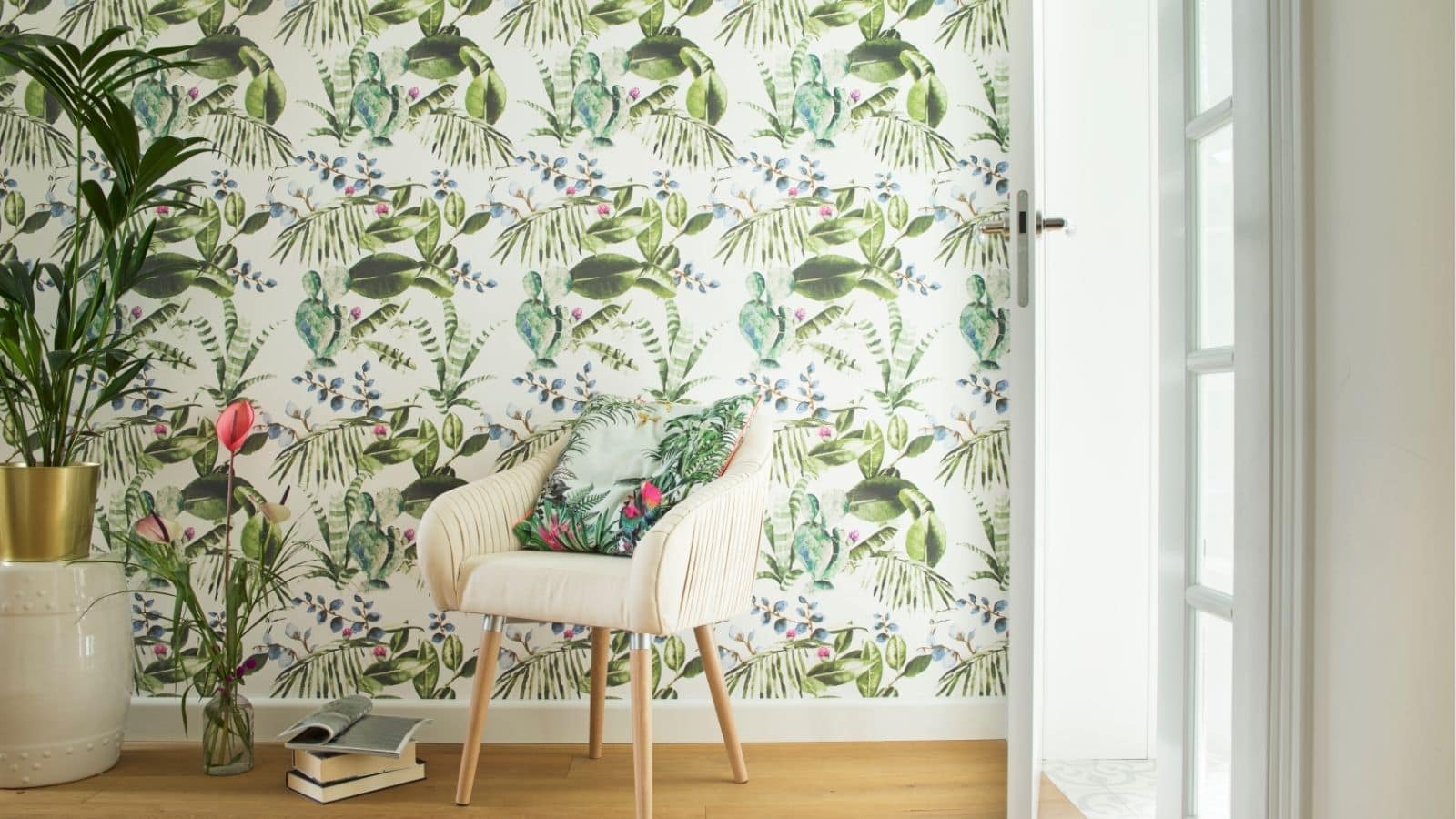 A cream-colored chair with a floral cushion sits by a tropical-themed wallpapered wall, next to a potted plant, books, and a glass door.