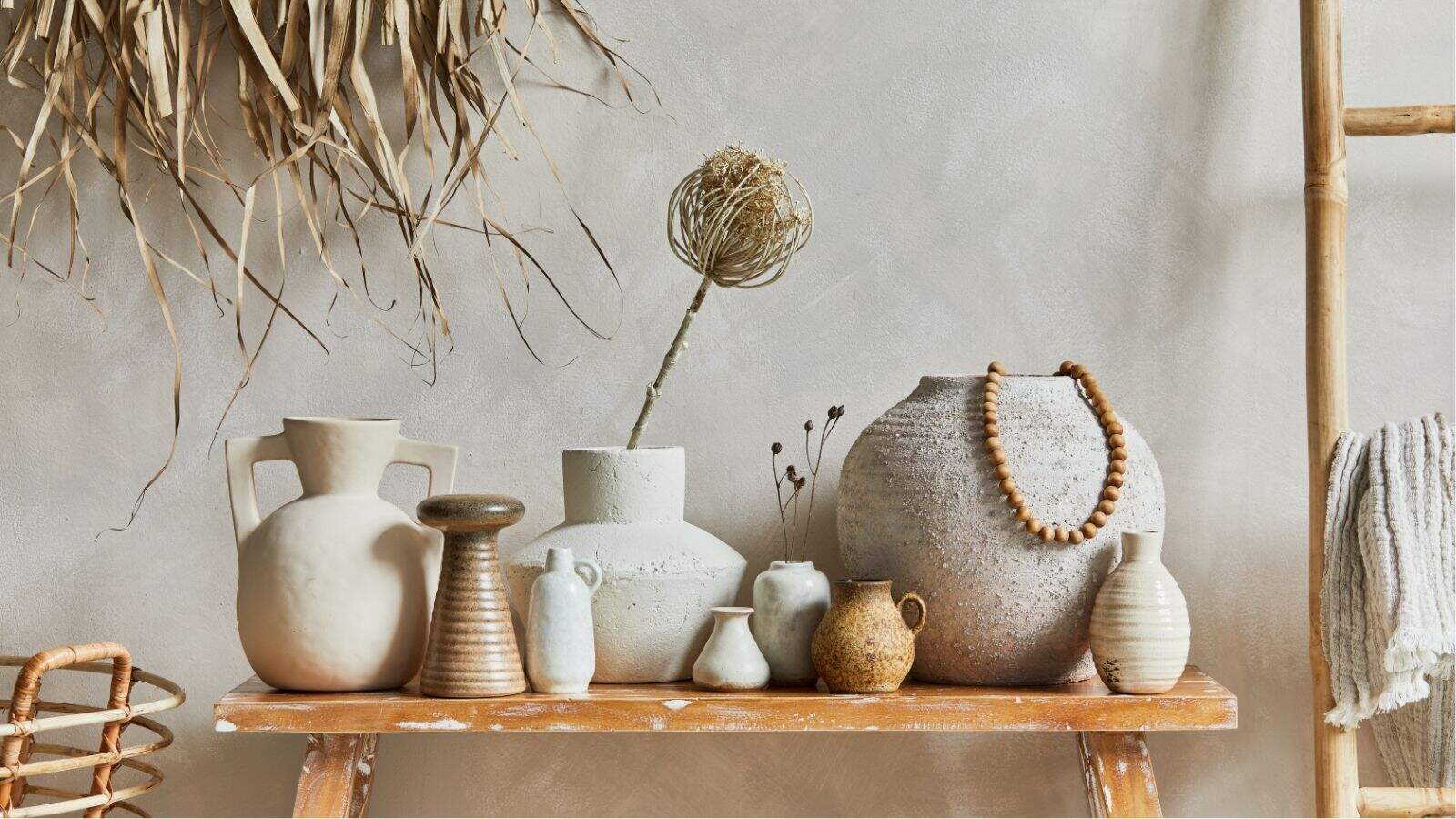 A wooden bench displays assorted neutral-toned ceramic vases and vessels, some with dried plants, against a beige wall with natural decor accents.