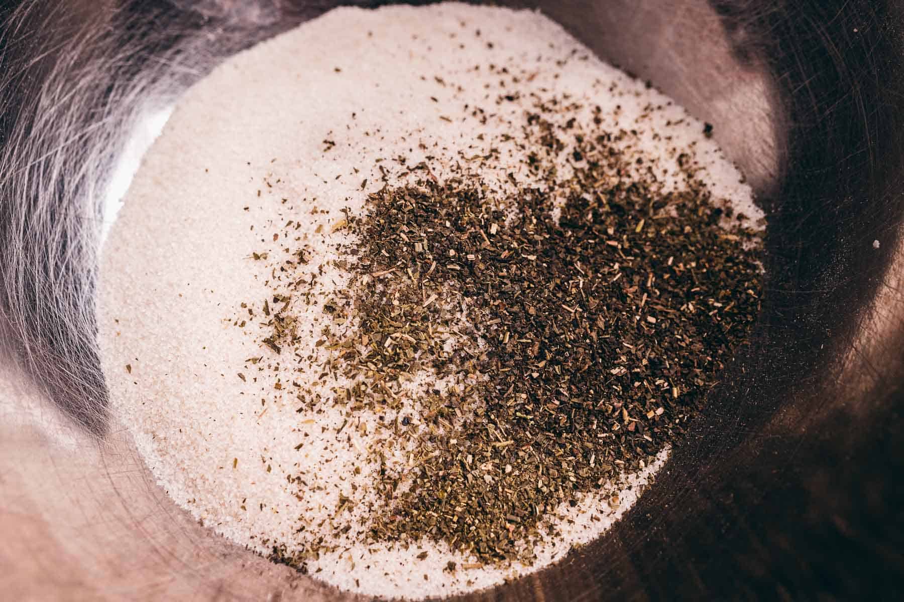 Close-up of granulated white sugar and dried green herbs, possibly basil or oregano, in a metal mixing bowl—perfect ingredients for creating a homemade green tea sugar scrub.