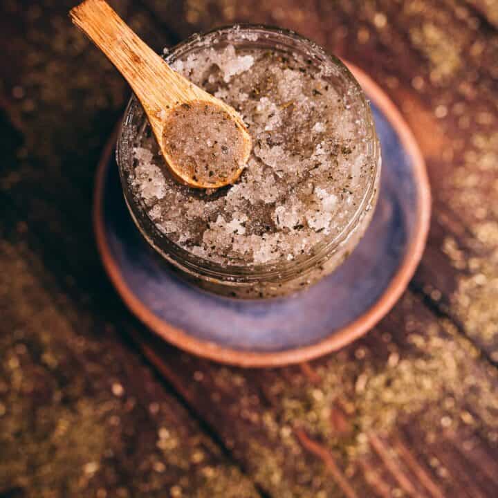 A jar of coarse salt mixed with herbs and green tea sugar scrub, with a small wooden spoon resting on top, placed on a ceramic plate on a rustic wooden surface.