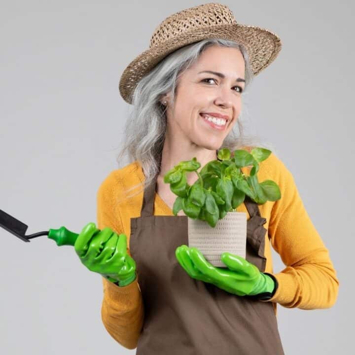 Woman wearing a straw hat and apron holds a potted plant and a small garden trowel, wearing green gloves, standing against a plain background.