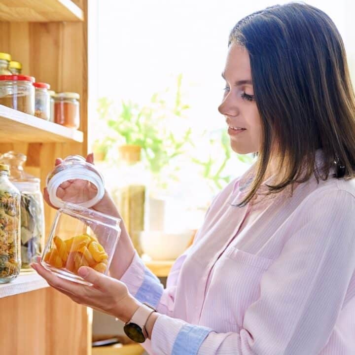 A woman in a light shirt places a jar of dried fruit on a wooden pantry shelf stocked with jars and food items.