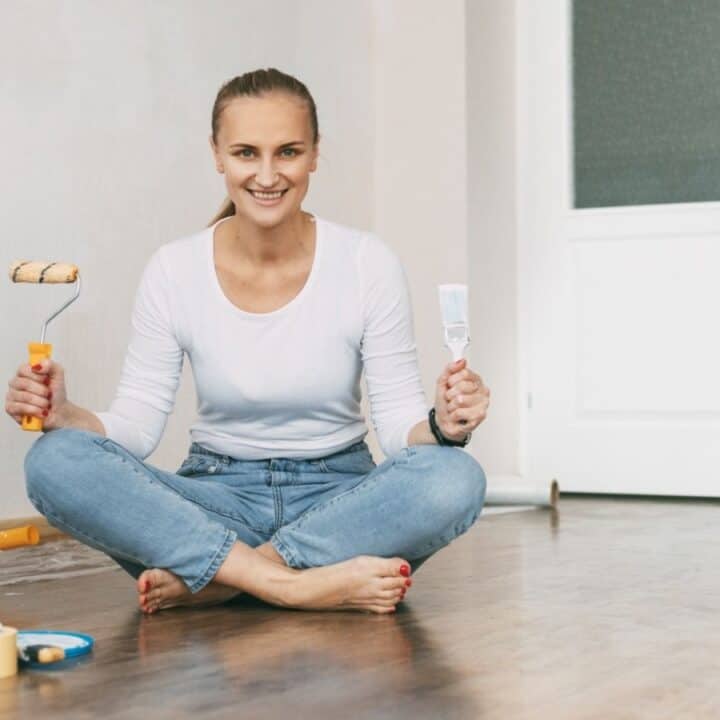 A woman sits cross-legged on a wood floor, smiling and holding a paint roller in one hand and a paintbrush in the other, with painting supplies nearby.