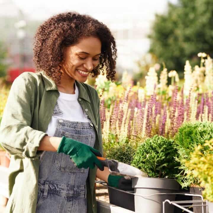 A woman wearing gloves and overalls tends to potted plants with a small trowel at an outdoor garden center.