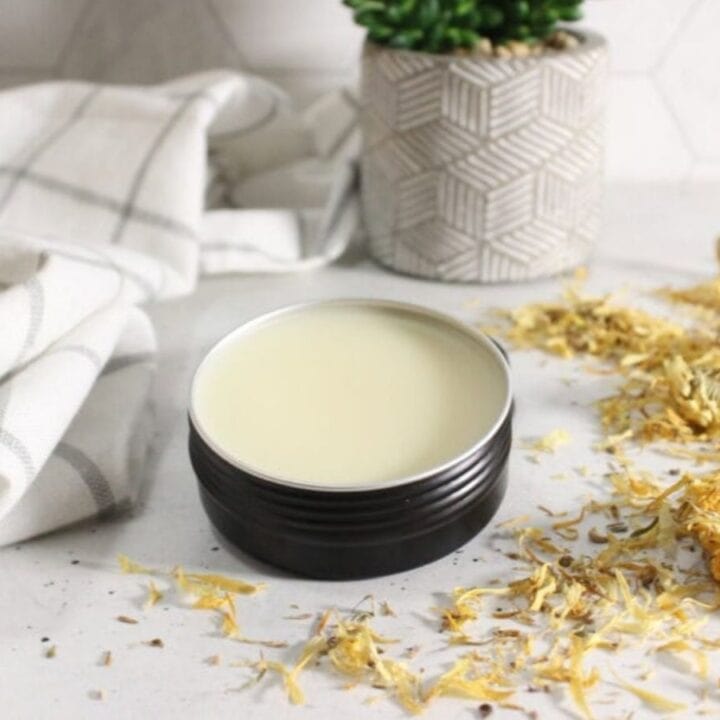 A metal tin filled with balm sits on a white surface next to dried yellow flowers, a folded cloth, and a small potted plant in the background.
