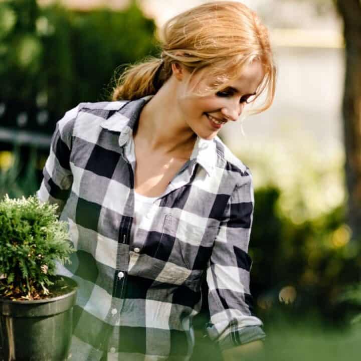 A woman in a plaid shirt holds a potted plant and smiles while gardening outdoors, surrounded by greenery.
