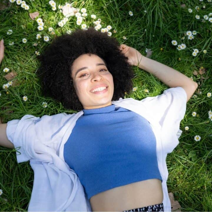 A person with curly hair lies on green grass dotted with small white flowers, smiling and looking up at the camera.