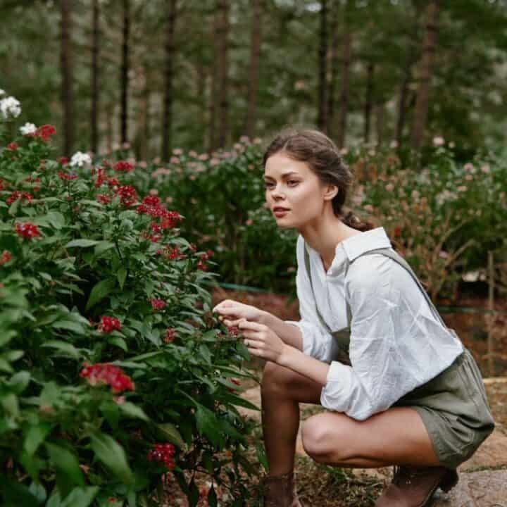 A woman in a white shirt and green shorts crouches next to a bush with red flowers in a garden surrounded by trees.