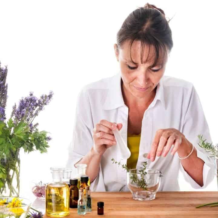 A woman in a white shirt is preparing herbal mixtures at a table with various fresh herbs, flowers, oils, and glass bowls.