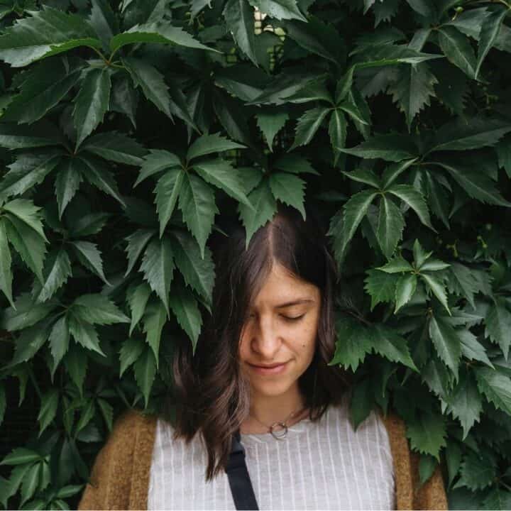 A woman with brown hair stands in front of a wall covered in large green leaves, looking down with a neutral expression.