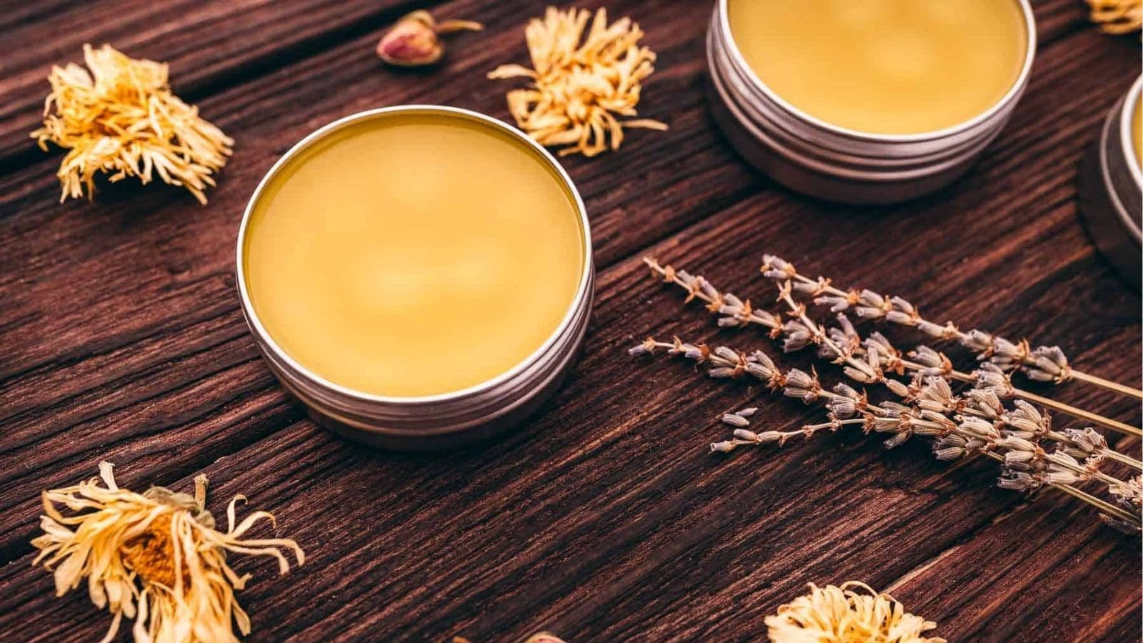 Two metal tins filled with yellow balm sit on a wooden surface, surrounded by dried flowers and lavender sprigs.