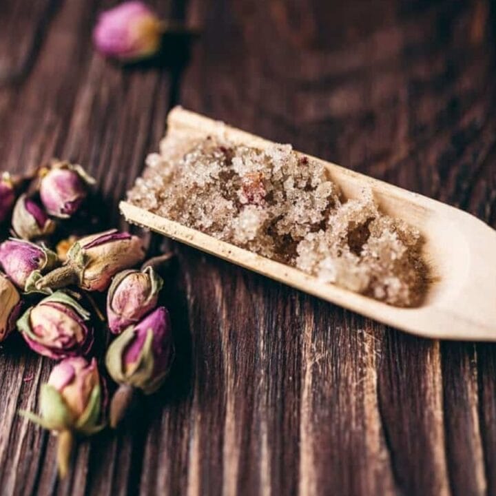 A wooden spoon holds coarse brown sugar next to several dried pink rosebuds on a dark wooden surface.