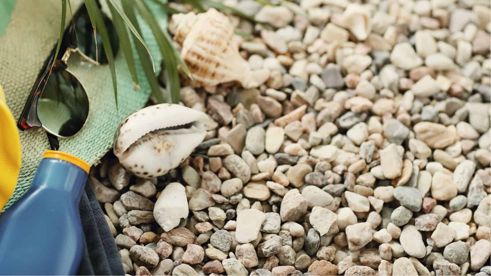 Sunglasses, sunscreen, and seashells rest on a textured surface of small, multicolored pebbles, with part of a green mat and some foliage visible.