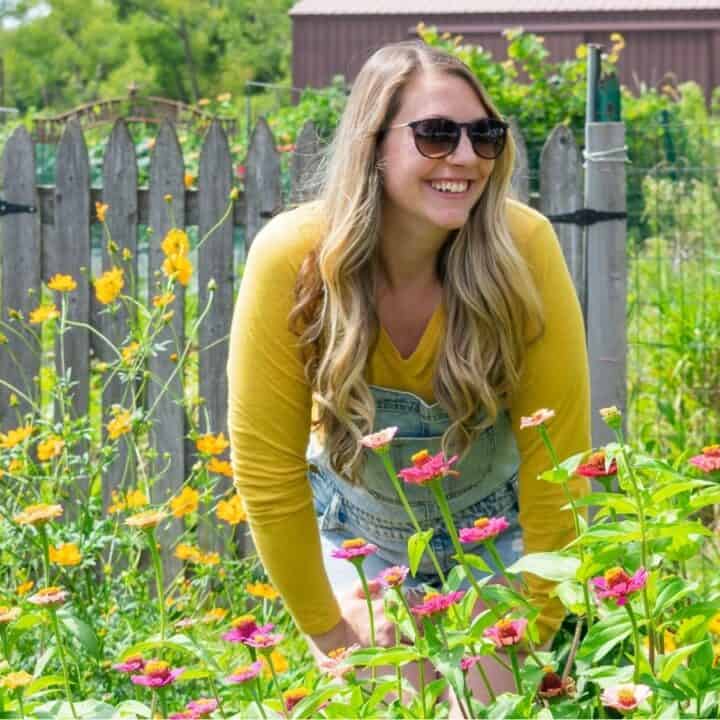 A woman wearing sunglasses, a yellow shirt, and denim overalls smiles while standing in a garden with blooming flowers and a wooden fence in the background.