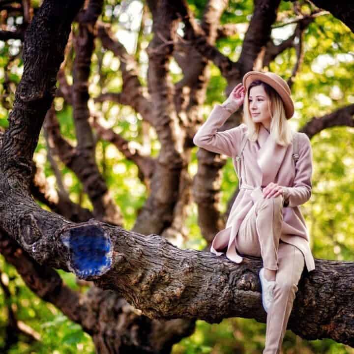 A person wearing a beige coat, hat, and boots sits on a thick tree branch in a forest, surrounded by green foliage.