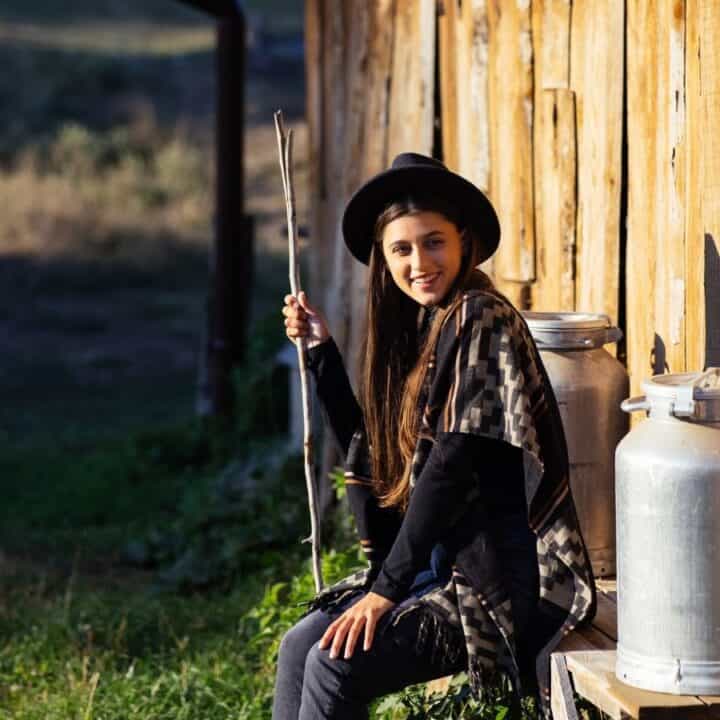 A person in a hat and patterned shawl sits on a bench outside a wooden building, holding a stick, with two metal milk cans beside them.