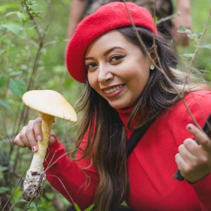 A woman in a red beret and sweater crouches in greenery, holding a yellow mushroom in one hand and a smartphone in the other, smiling at the camera.